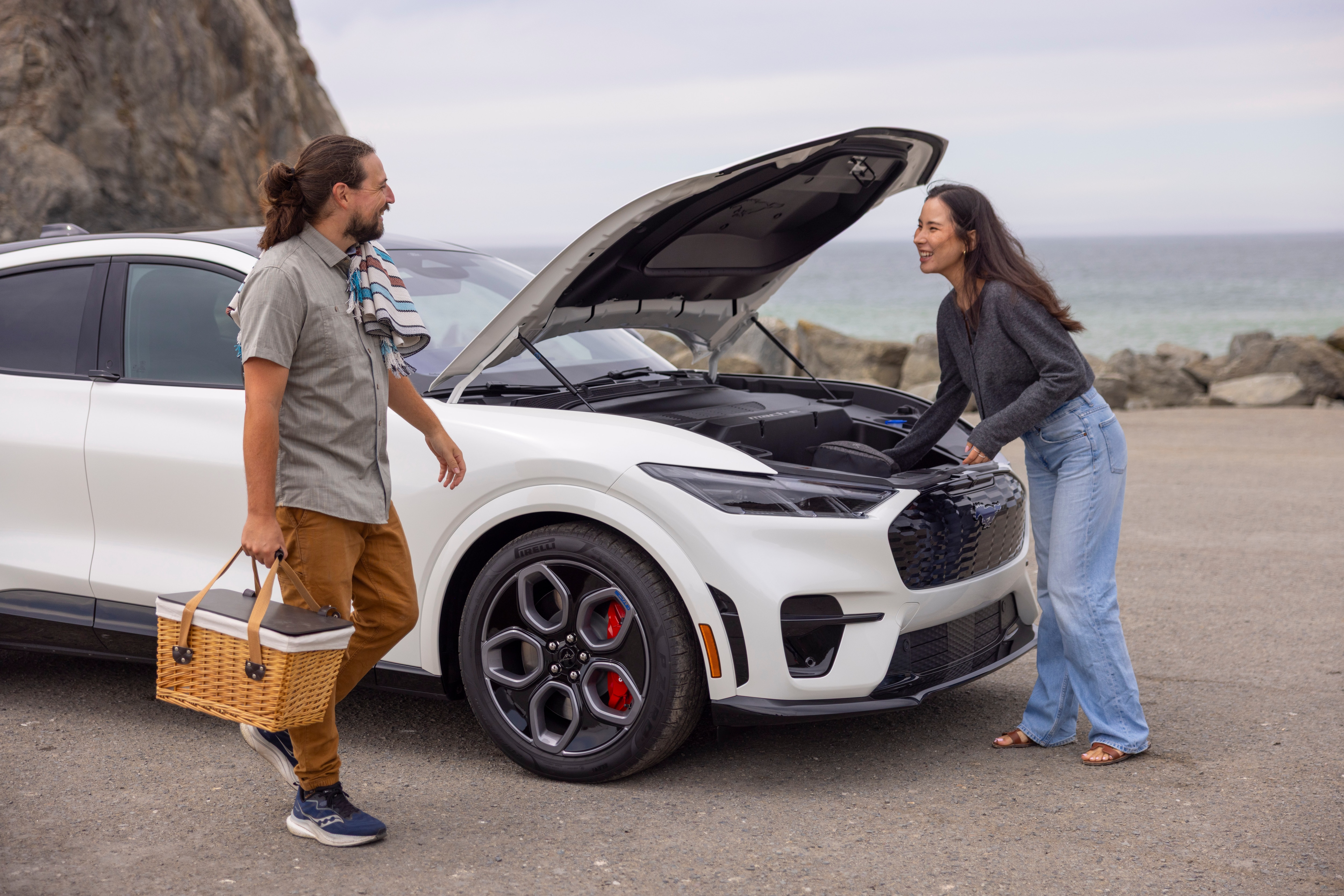 A man and woman stand on the beach holding a picnic basket near a white Mustang Mach-E.